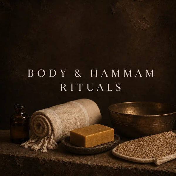 A styled spa setup with a rolled cream towel, a rectangular bar of golden soap on a dark stone dish, a small amber bottle, a brass bowl, and a woven mat, set against a dark brown textured background with the text 'BODY & HAMMAM RITUALS'.