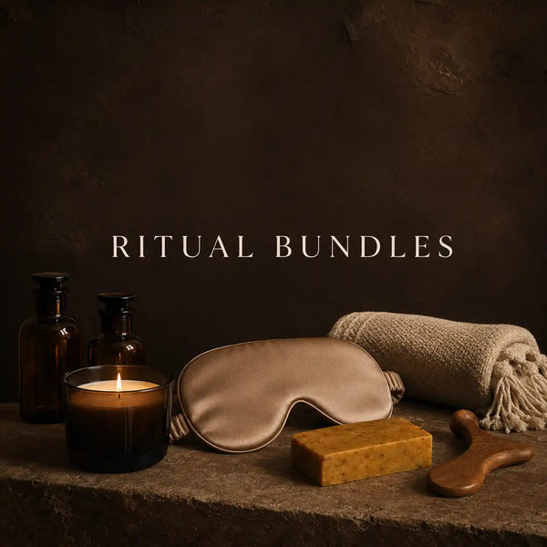 A styled spa ritual bundle set on a stone surface against a dark brown textured background, featuring a satin sleep mask, a lit candle in a dark amber glass holder, two amber bottles, a folded beige towel, a yellow soap bar, and a wooden massage tool. The text 'RITUAL BUNDLES' is centered above the items.